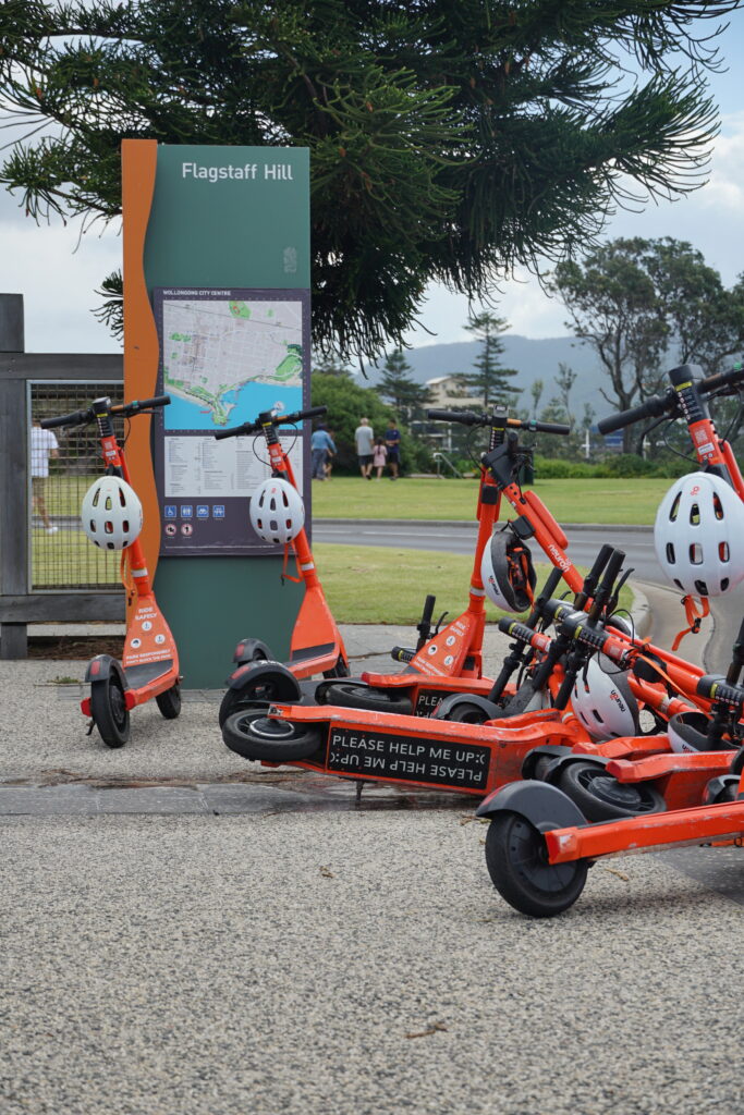 A number of e-scooters lined up at Flagstaff Hill, with a few knocked over, revealing "Please help me up :(" written underneath.
