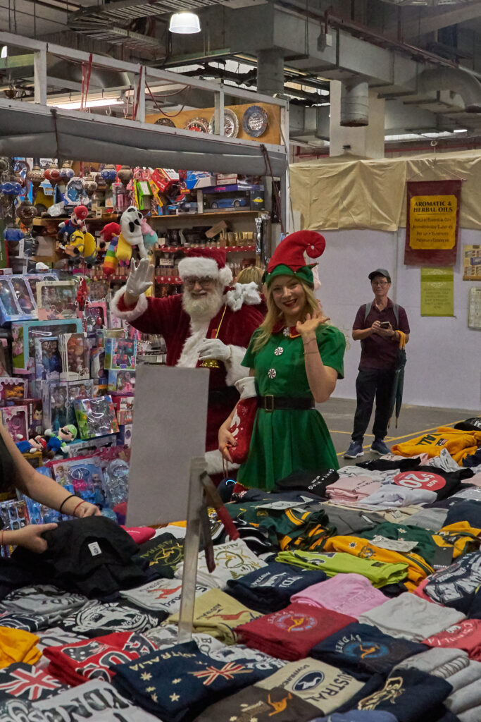 Two people, dressed as Santa Claus and a helper elf, walking past stalls in Market City.