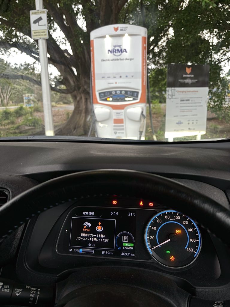 A picture from within a car with the dash visible, indicating a charge of 15% remaining and an estimated range of 39km. Beyond the windscreen is an electric vehicle charger.