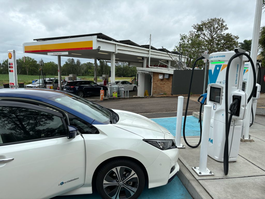 A Nissan Leaf parked in front of one electric vehicle charger, with another unoccupied charger behind. In the background is a Shell petrol station.