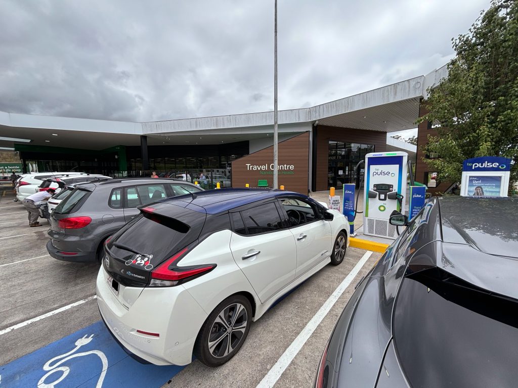 A row of cars parked outside BP Nambucca Heads, with a Nissan Leaf in the foreground connected to an electric vehicle charger.