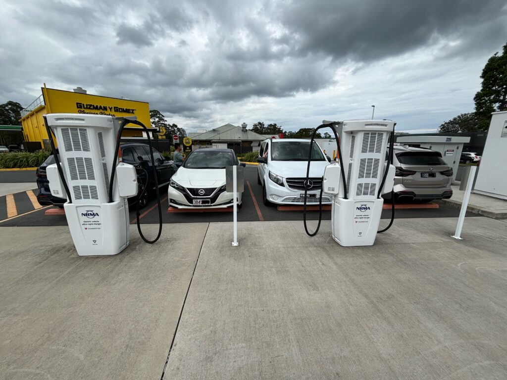 A row of four cars in the background, and two electric vehicle chargers in the foreground with the back of the units facing towards the camera.