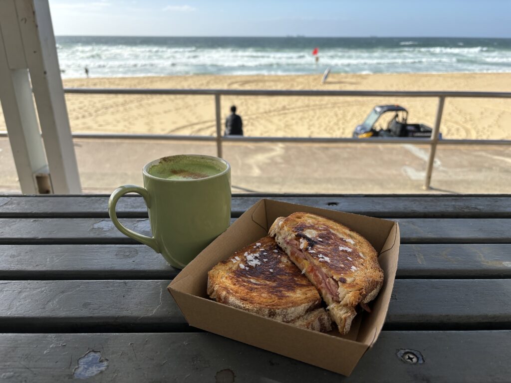 A green mug and toasted sandwich sit on a bench overlooking Newcastle Beach.