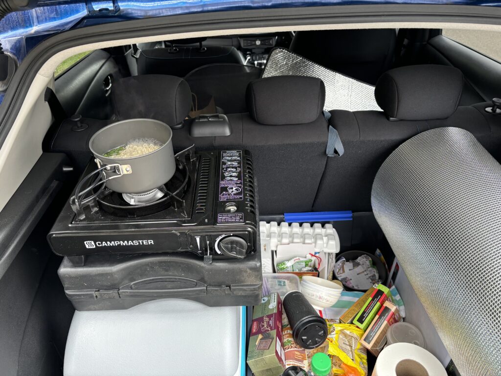 The boot of a car with a camping stove on top of its own carry box on a closed cool box, next to the cool box is a large pile of food and utensils. The camping stove is lit, with a small pot on top and instant noodles inside boiling.