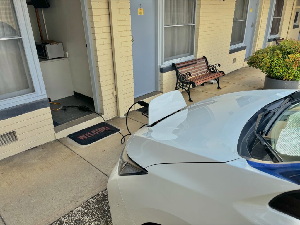 A Nissan Leaf parked close to the door of a motel room, with an EVSE connected via a red/yellow extension lead from the room.