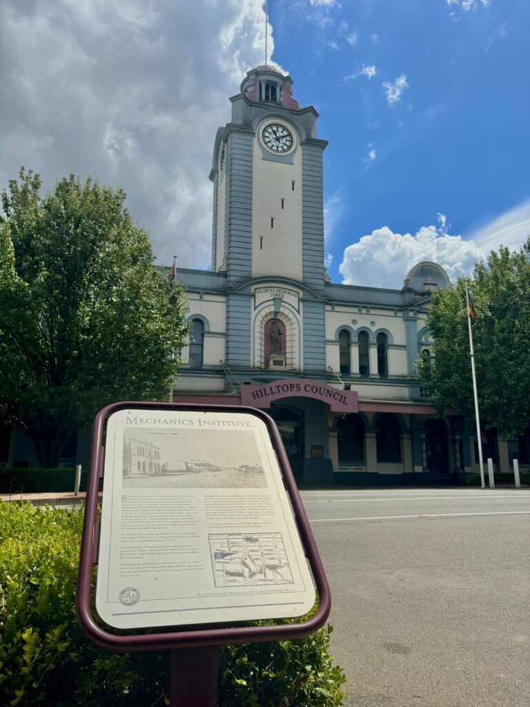 A building with a tall clock tower, with an information plaque in the foreground describing its history from across the road.