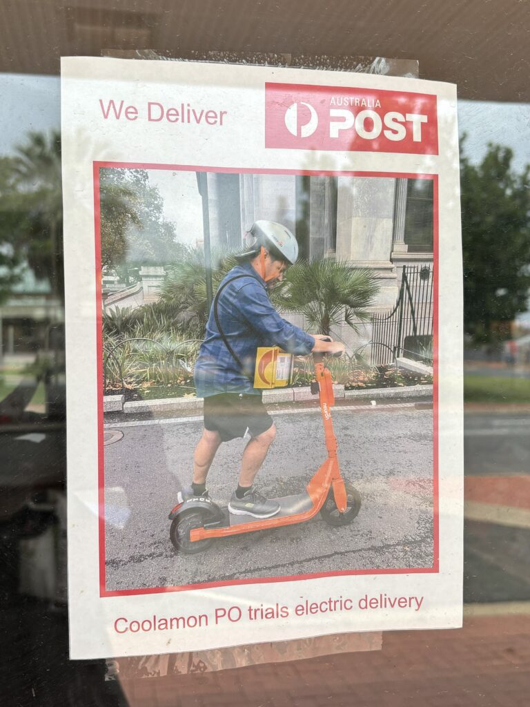 A poster stuck on a window with the Australia Post logo that reads: "We Deliver/Coolamon PO trials electric delivery" and an image of a person on an orange electric scooter and an two packages strapped on their shoulder.