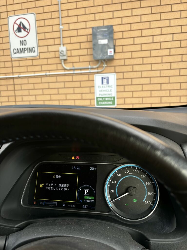 A photo from inside the driver's position in a car, with a brick wall and electric vehicle charger in the background. The instrument panel in the car displays the warning: "バッテリー残量低下　充電してください", with the battery reading 3% charge and an estimated range of "---" kilometres.